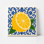A square decorative tile or print featuring a large yellow lemon with green leaves on a cream background, surrounded by a blue floral scroll pattern, shown against a light gray wall.