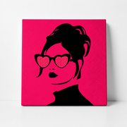 A square canvas wall art print featuring a stylized black silhouette of a woman with heart-shaped sunglasses on a bright hot pink background, shown leaning against a white wall.